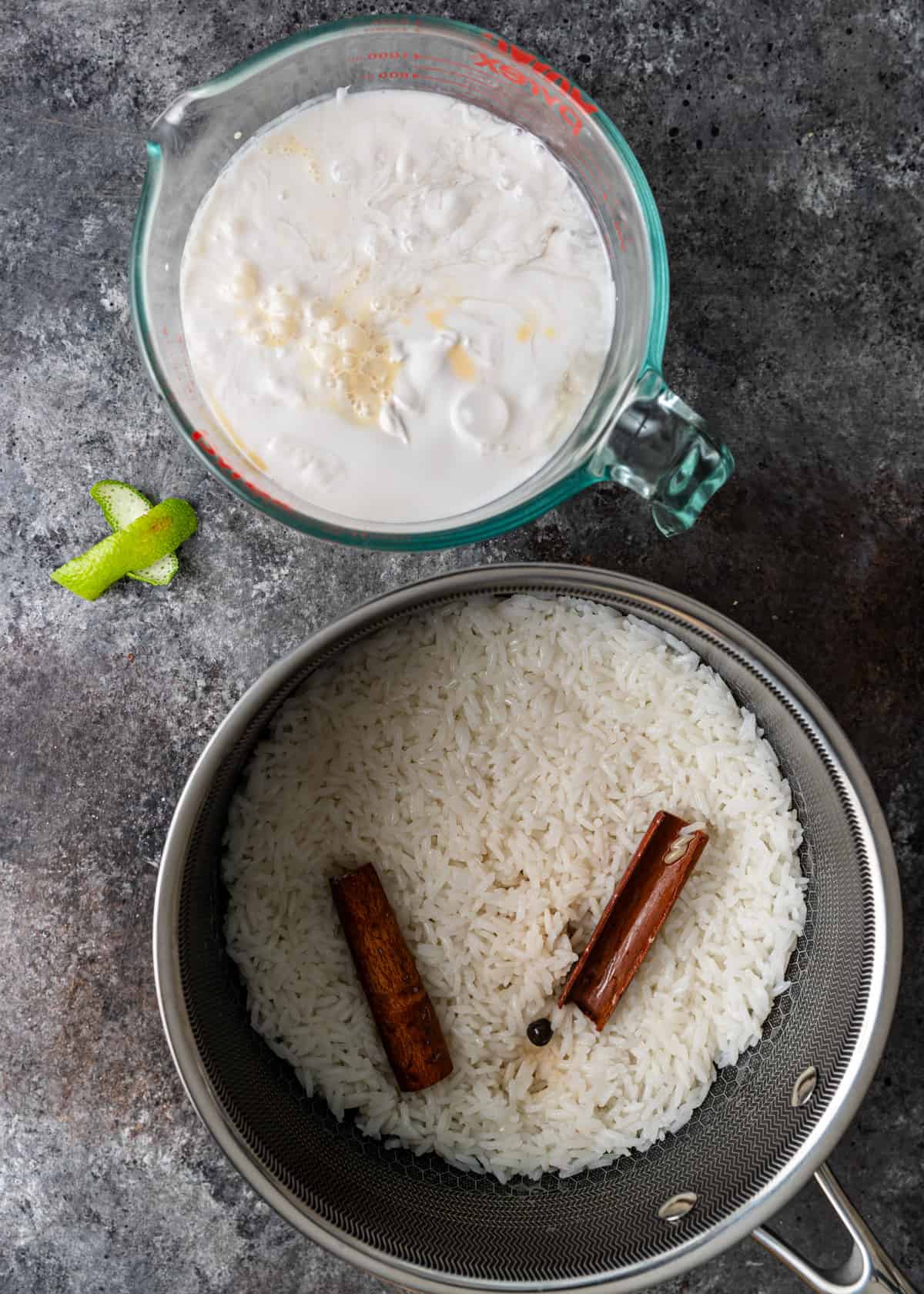 overhead: cooked rice in a saucepan with cinnamon sticks and a glass measuring cup full of milk to the side
