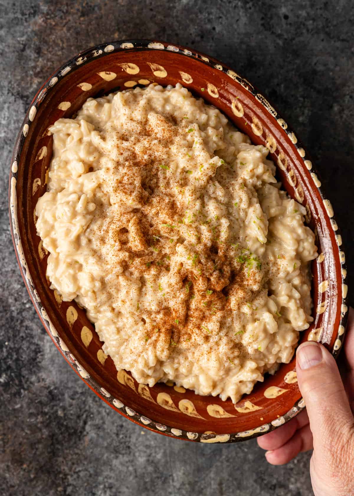 overhead: a hand holding a ceramic serving tray of Mexican rice pudding with allspice on top