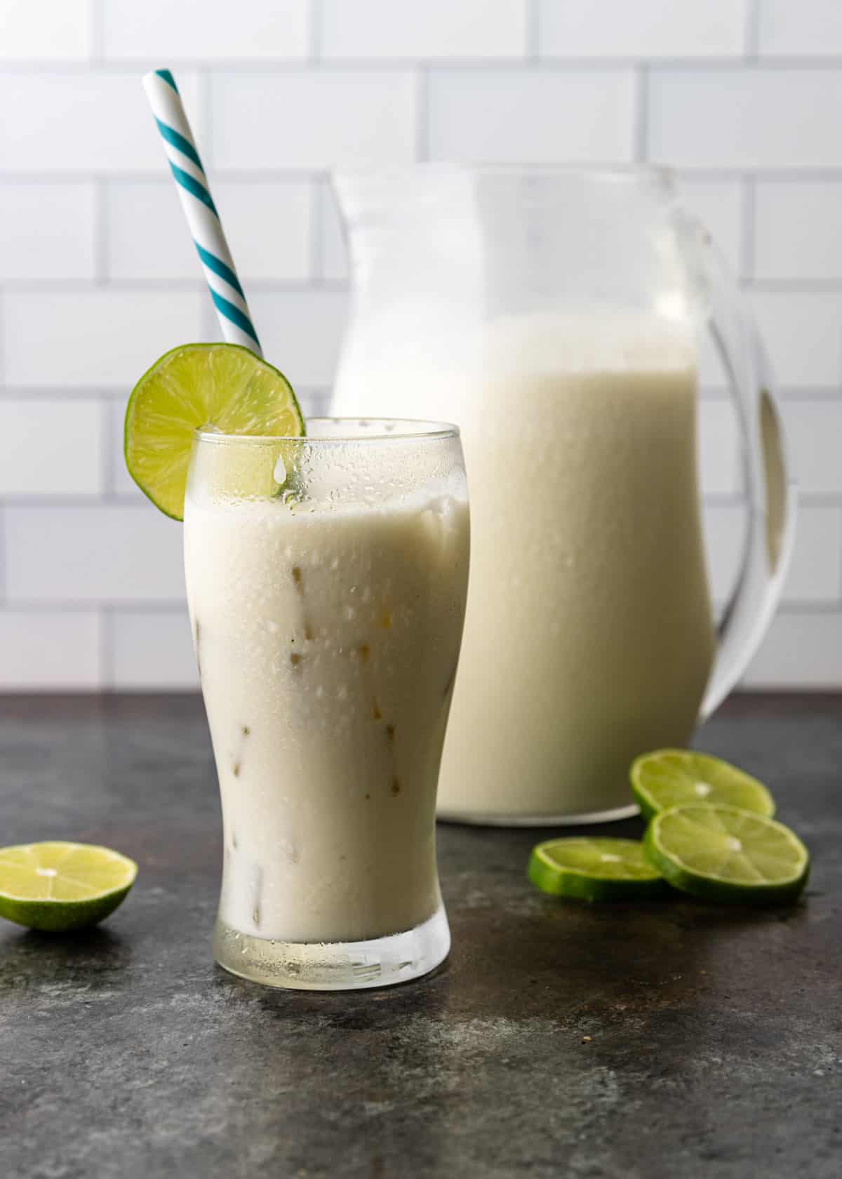 a tall glass of cold Brazilian lemonade with a lime round and blue and white straw. A pitcher of lemonade is visible in the background