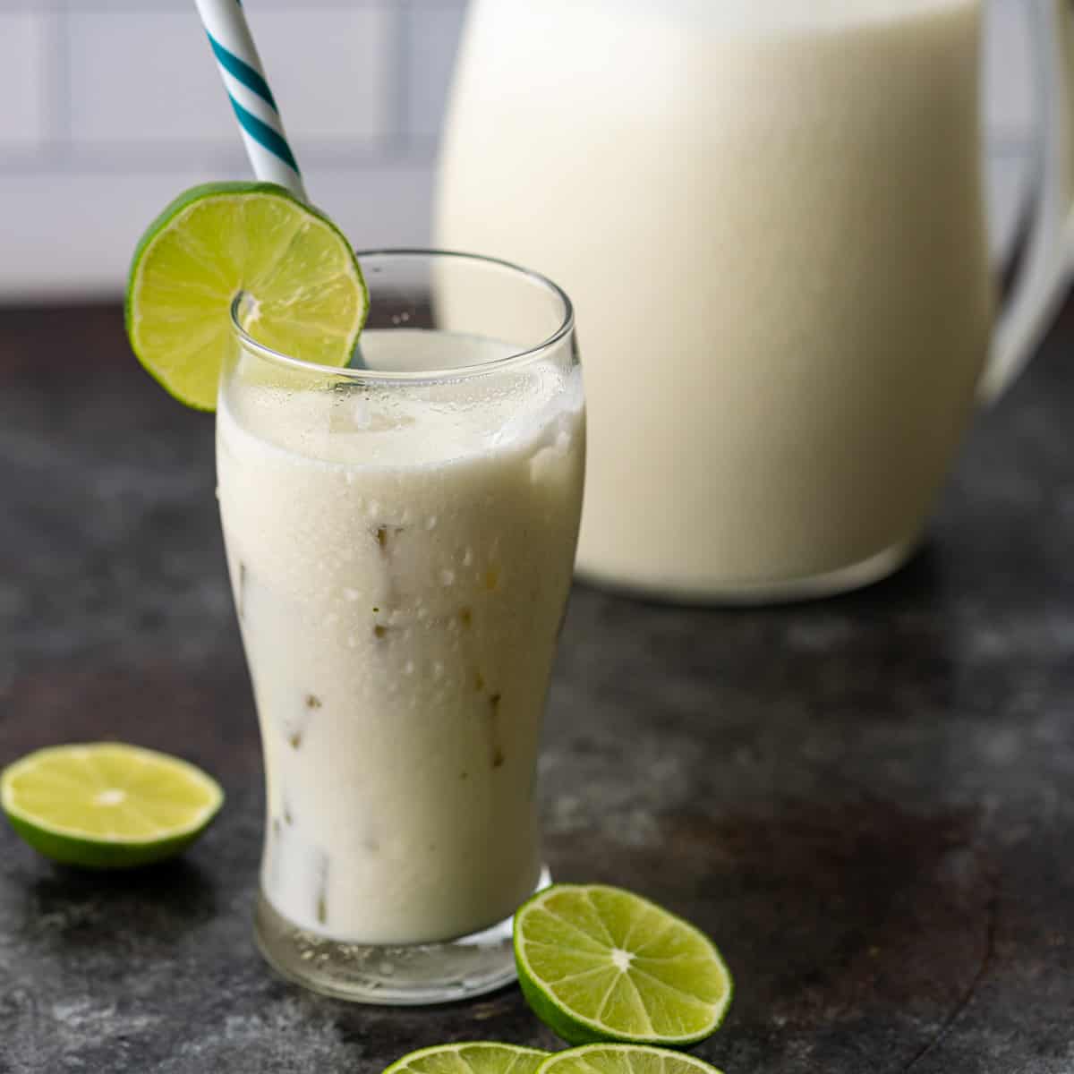 a tall glass of cold Brazilian lemonade with a lime round and a blue and white straw. A pitcher of lemonade is visible in the background