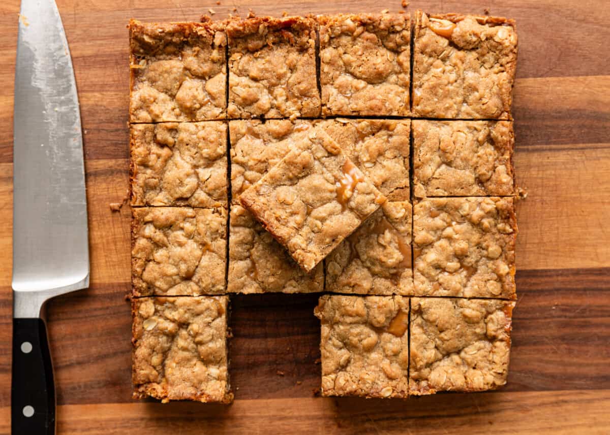overhead: caramel oatmeal bars sliced on a cutting board with a piece taken out and a large knife to the left