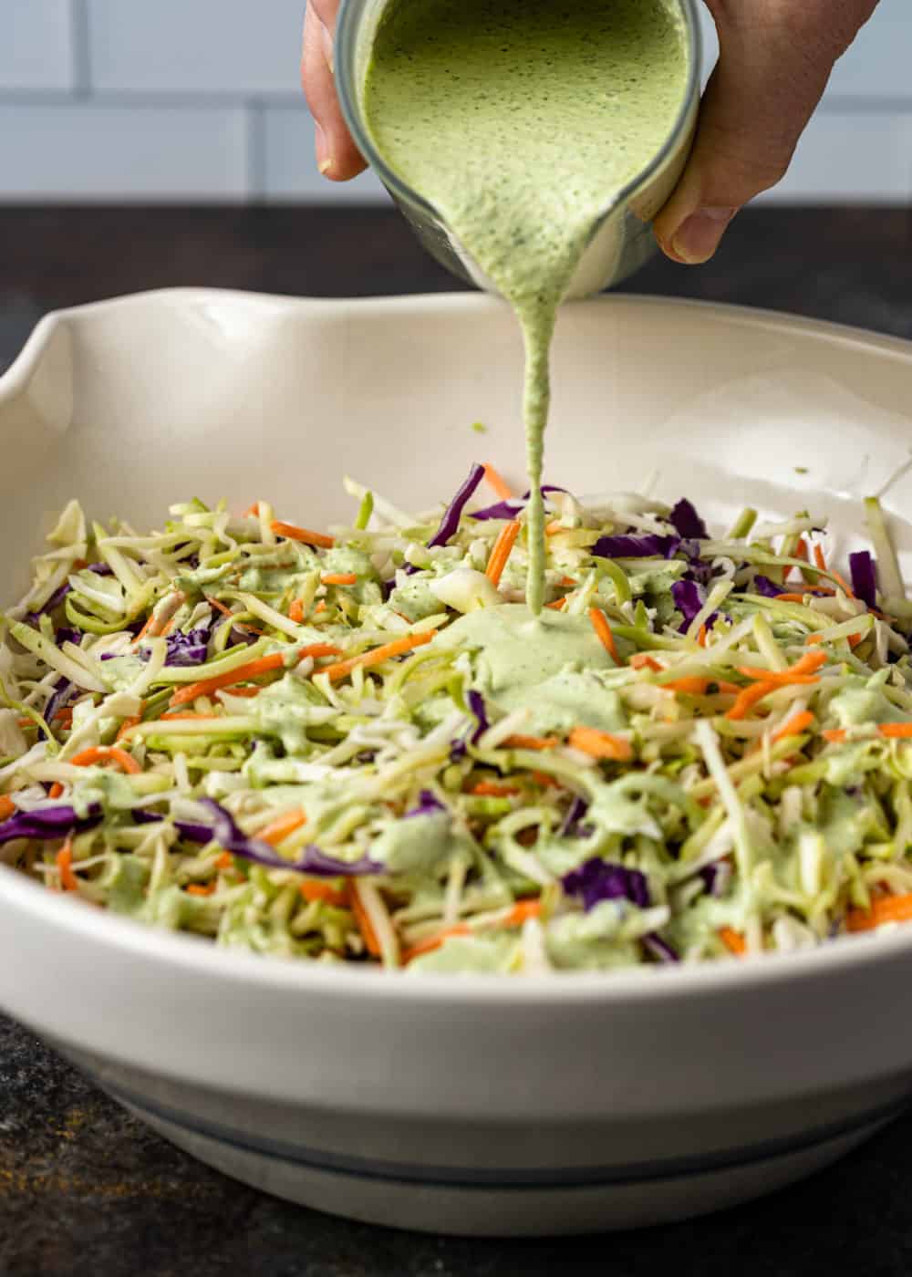 making a spicy coleslaw recipe by pouring creamy green dressing over bowl of shredded cabbage and carrots