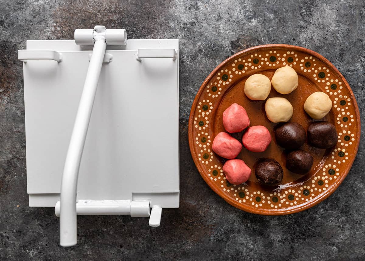 A closed tortilla press with a plate of conchas dough balls to the right