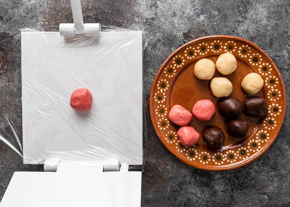 An opened tortilla press with a pink pan dolce in the center and a plate of conchas dough balls to the right