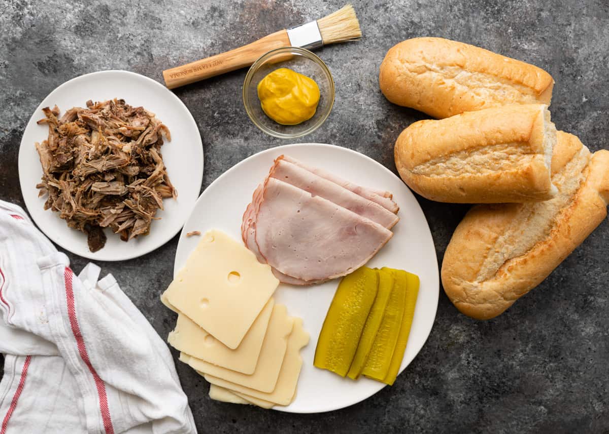 Ingredients for Cuban sandwiches including pork, mustard, ham, pickles, Swiss cheese and sliced bread.