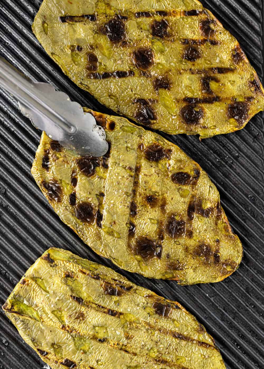 overhead image: 3 cactus paddles on a grill, one being turned with metal tongs