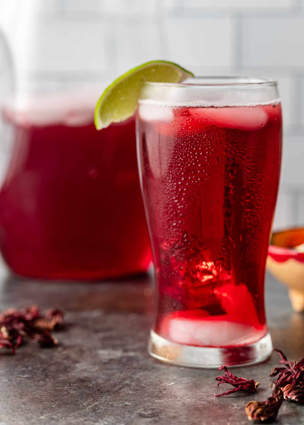 a tall glass of agua de jamaica with ice and a lime slice garnish with dried hibiscus flowers on counter