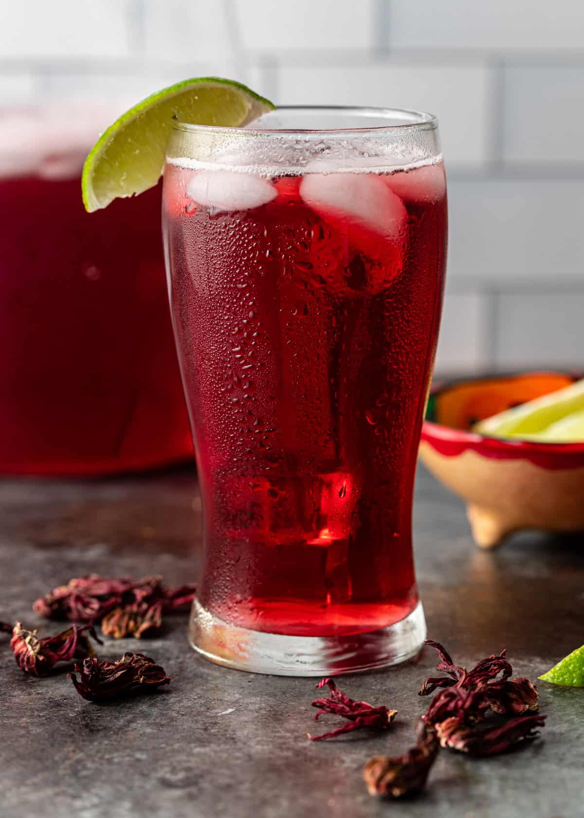 a tall glass of jamaica drink with ice and a lime slice as a garnish with dried hibiscus flowers on counter