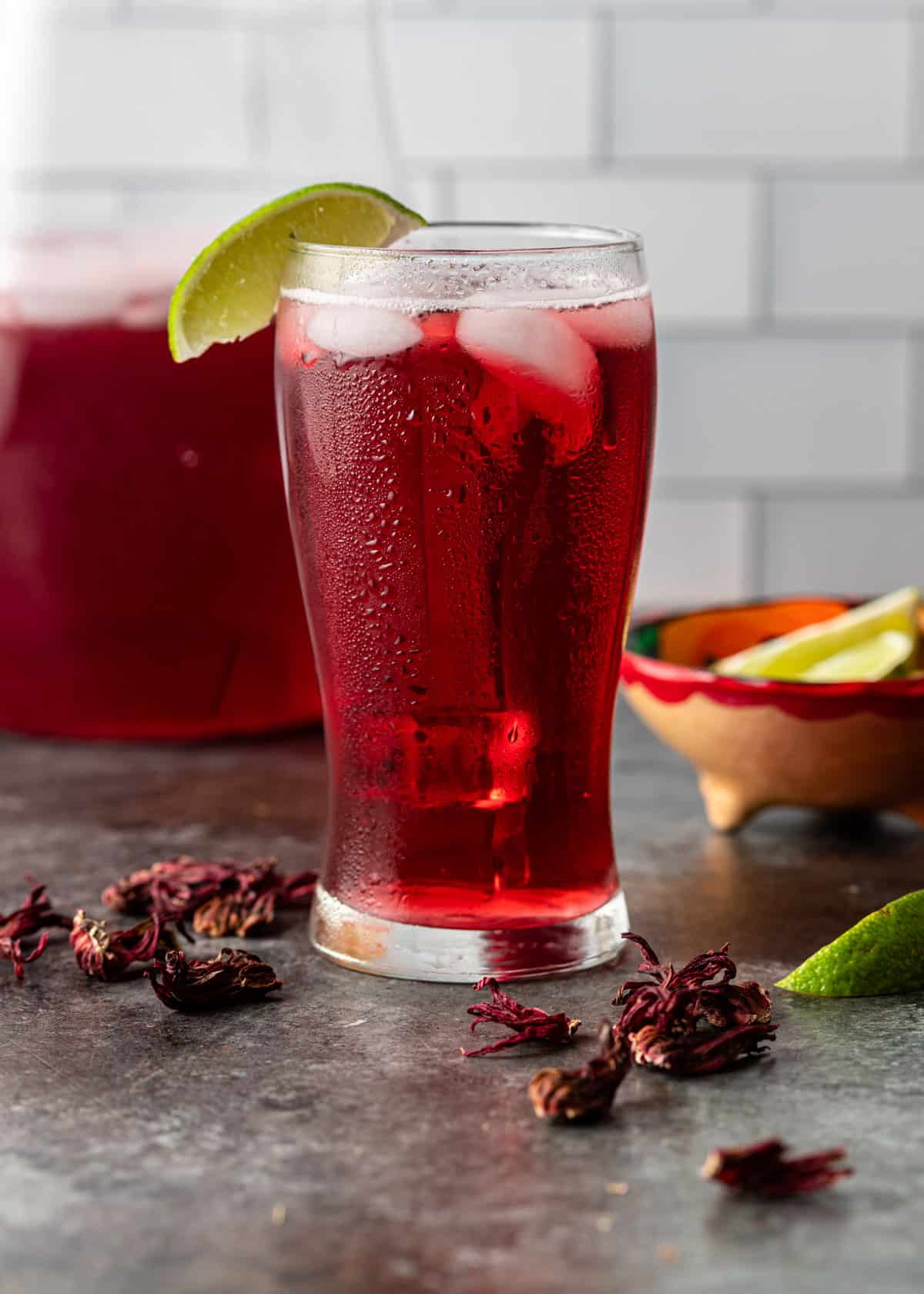 a tall glass of hibiscus iced tea garnished with a lime slice with dried hibiscus flowers on counter