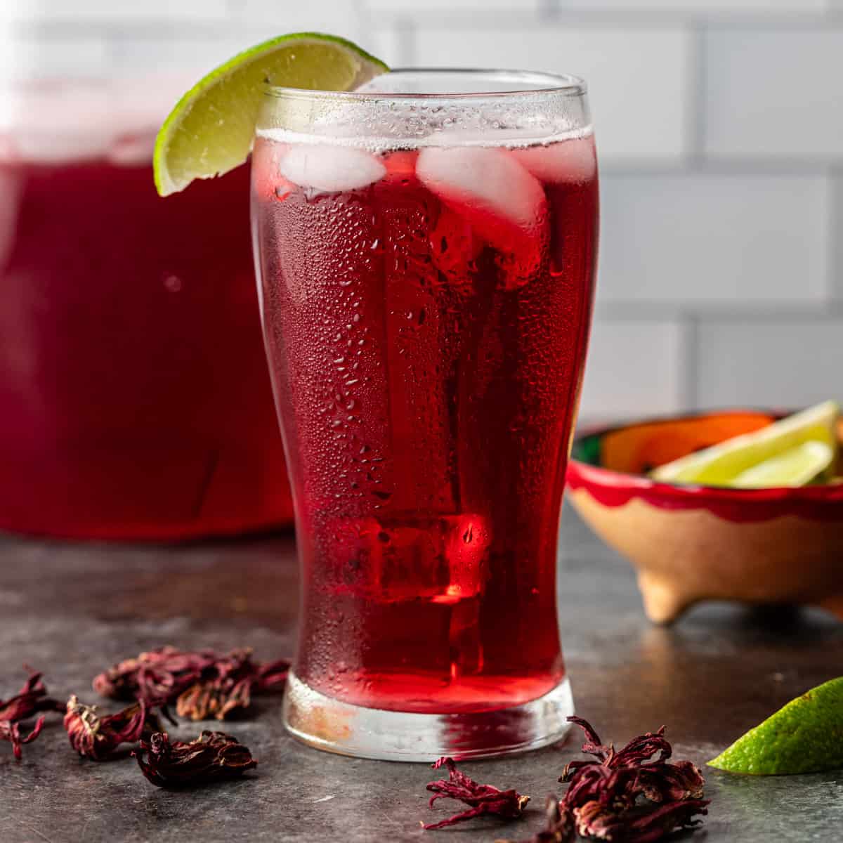 a tall glass of hibiscus iced tea with ice and a lime slice as a garnish with dried hibiscus flowers on counter 