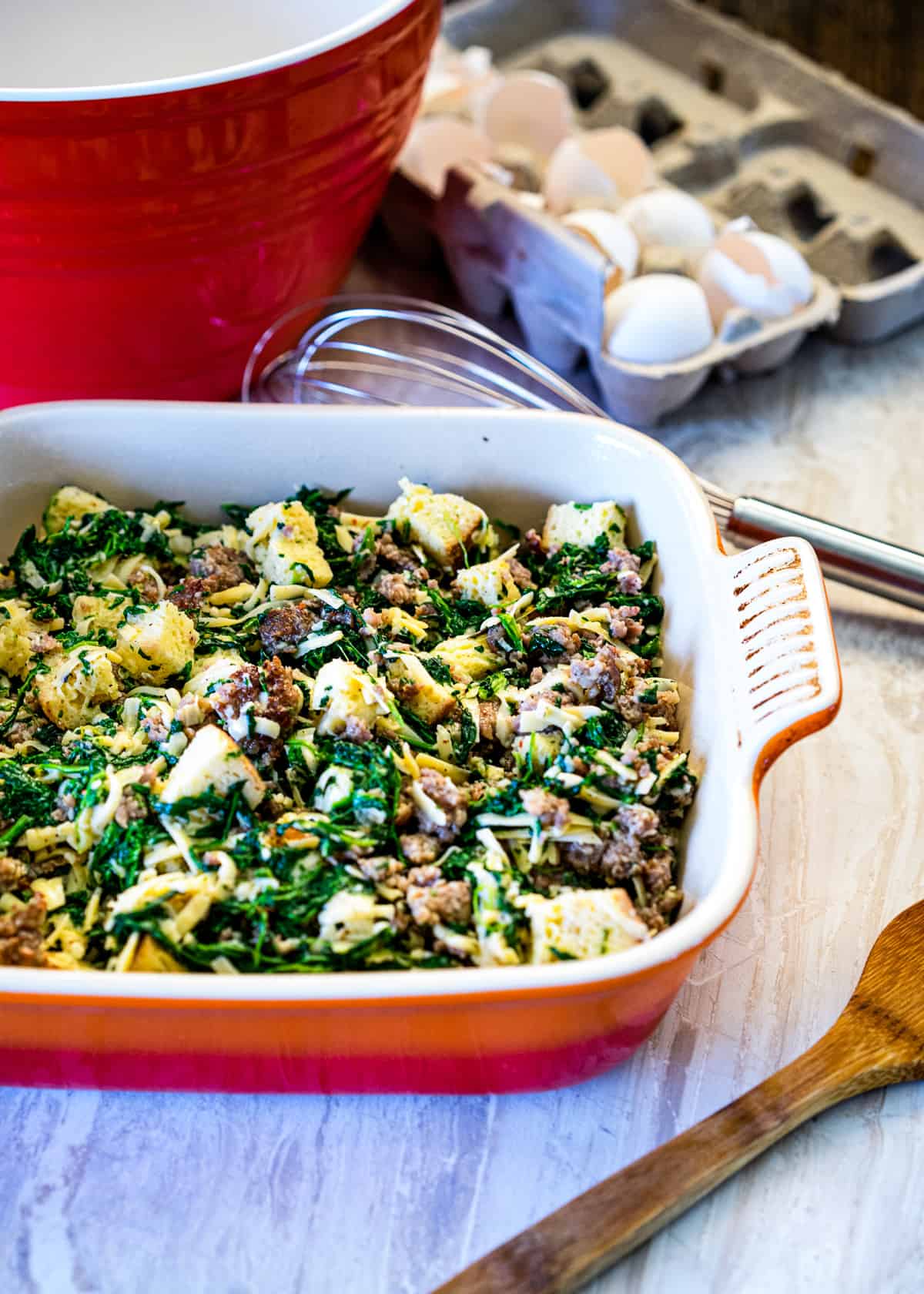 carton of fresh eggs next to red mixing bowl, whisk, and a baked eggs casserole