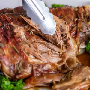 close up of tongs pulling tender meat from a roast leg of lamb