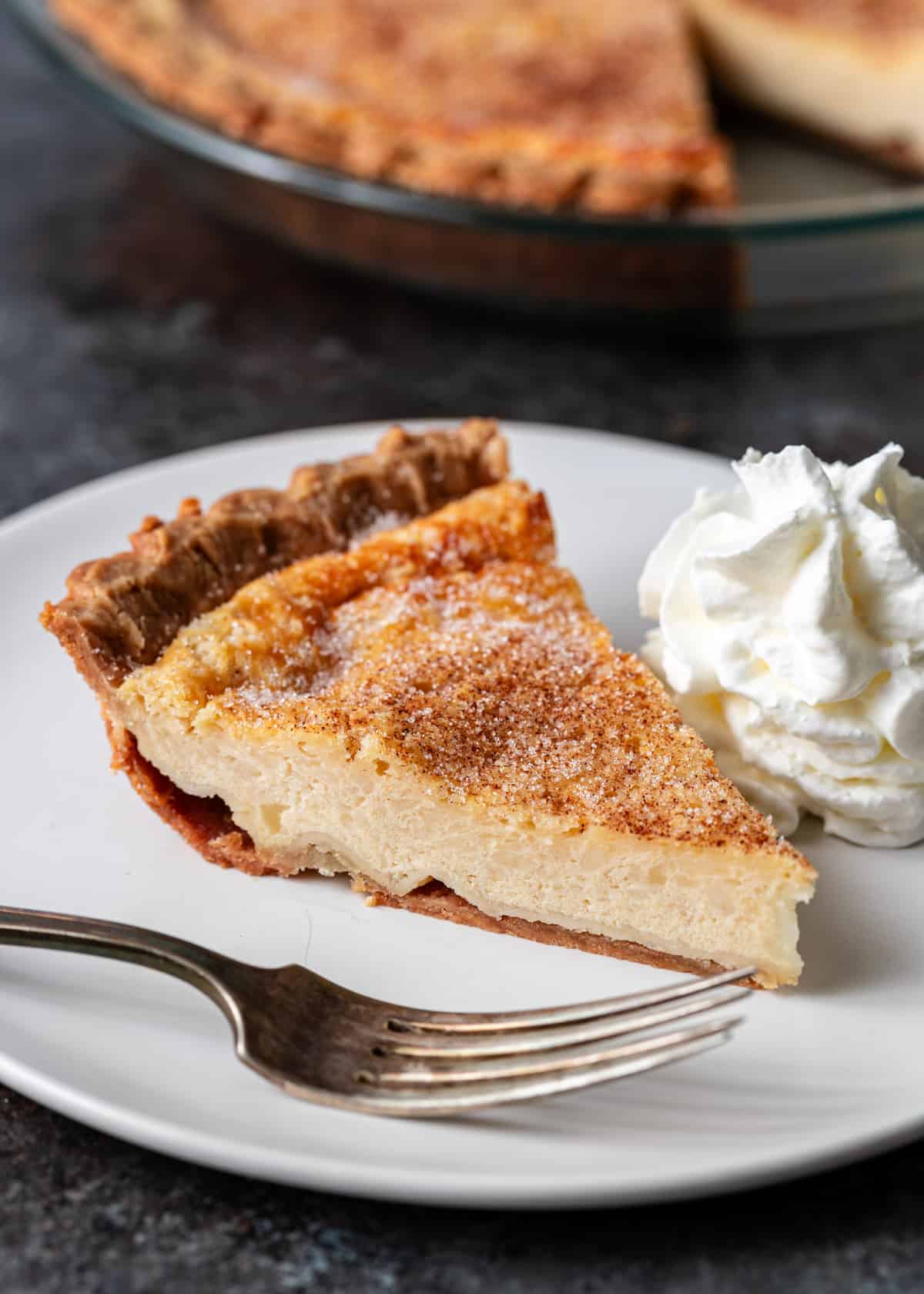 a slice of buttermilk pie on a white plate with whipped cream and a fork on either side