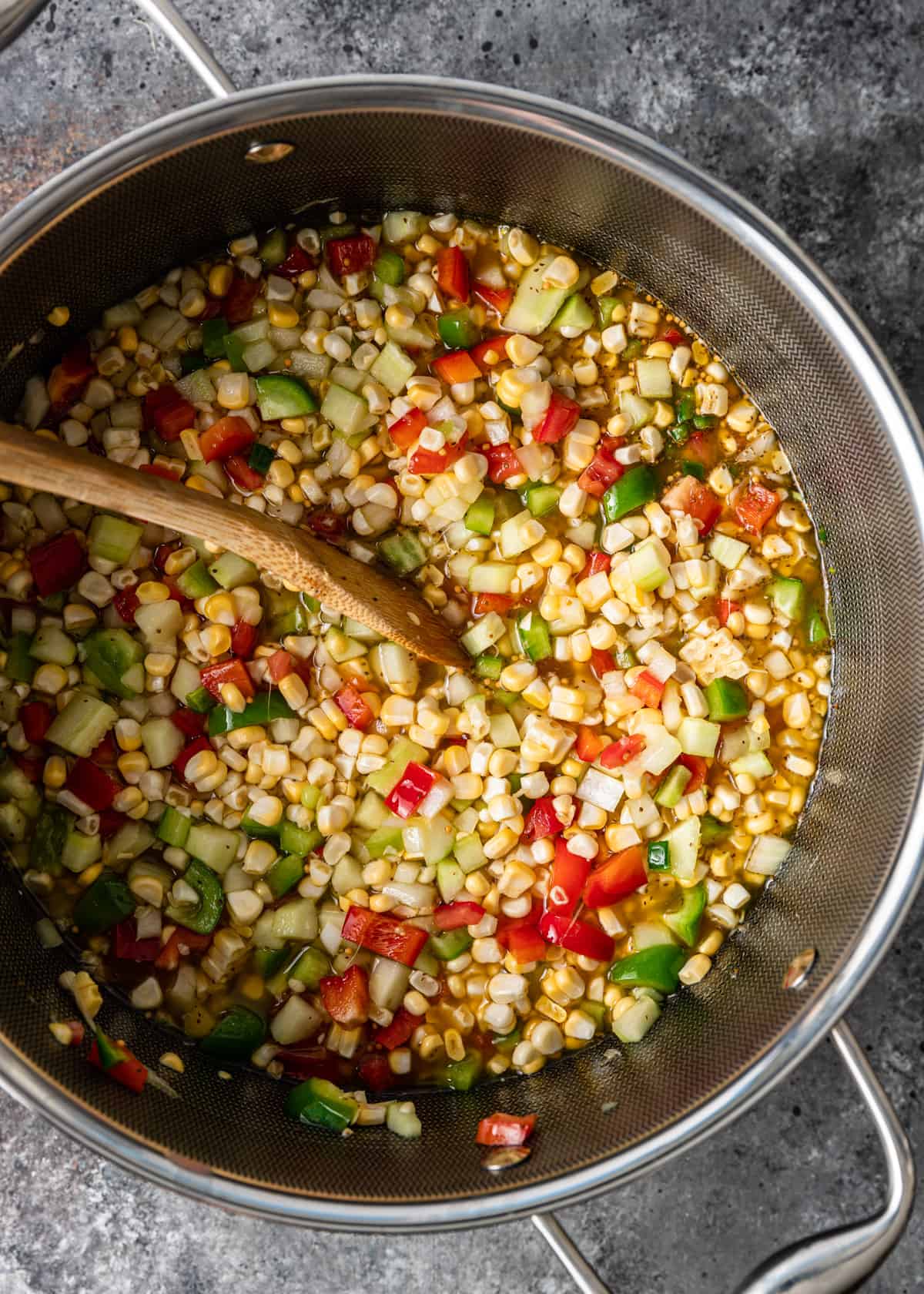 overhead: corn, tomatoes, onion, cucumber, and peppers in a large stock pot with a wooden spoon